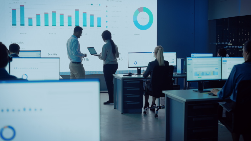 Two Financial Data Analysts Having a Meeting in a Modern Monitoring Office with Analytics Feed on a Big Digital Screen. Monitoring Room with Brokers and Finance Specialists Sit in Front of Computers. Royalty-Free Stock Footage #1056387980