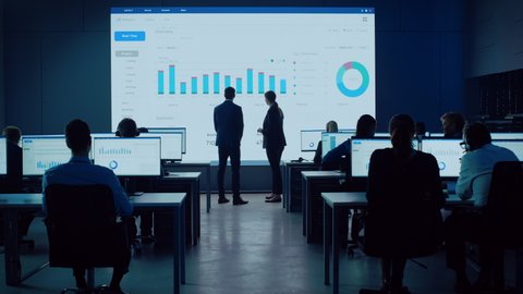 Two Financial Data Analysts Having a Meeting in a Modern Monitoring Office with Analytics Feed on a Big Digital Screen. Monitoring Room with Brokers and Finance Specialists Sit in Front of Computers.