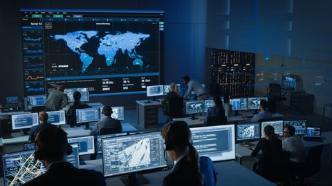 Government Officers Work on a Computers with Surveillance CCTV Video in a Police Monitoring Center with Global Map Tracking on a Big Digital Screen. Employees Sit in Front of Displays with Big Data