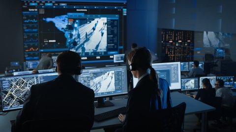 Female Officer Works on a Computer with Surveillance CCTV Video Footage in a Police Monitoring Center with Global Map Tracking on a Big Digital Screen. Employees Sit in Front of Displays with Big Data