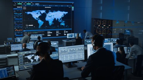 Moving Shot of Officers in a Surveillance Control Center with Police Global Map Tracking on a Big Digital Screen. Monitoring Room Employees Sit in Front of Computer Displays and Analyze Big Data.