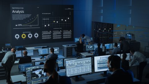 Static Shot of Professional Traders Working in a Modern Monitoring Office with Live Analytics Feed on a Big Digital Screen. Monitoring Room Brokers and Finance Specialists Sit in Front of Computers.