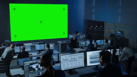 Big Green Screen Horizontal Mock Up in a Control Center Room with Engineers and Controllers Working on Computers. Team of Telecommunication Employees Work in Monitoring Room Full of Displays.