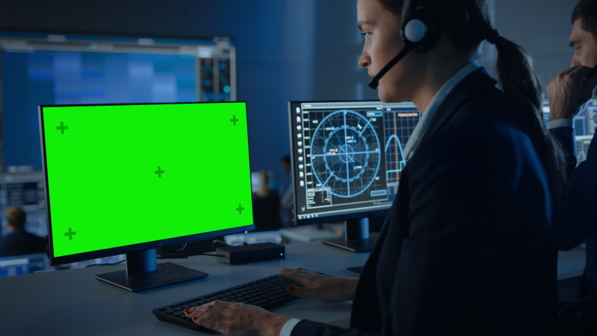Green Screen Horizontal Mock Up on a Computer Display Used by a Female Controller in a Mission Control Center Room with Other Scientists. Team of Engineers Work in Monitoring Room Full of Displays.