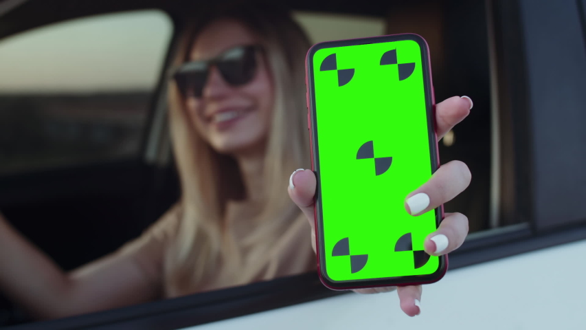 A happy girl is sitting behind a car, holding a phone with a chromakey in camera focus. Cute girl shows a phone with a green screen from the car Royalty-Free Stock Footage #1056388205