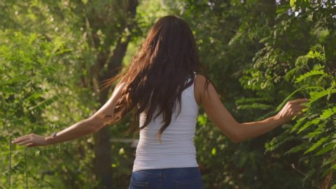 Tanned young woman with sexy lips and curly hair enjoys nature in summer forest. Beautiful brunette smiles and walks fast among green trees in sunlight. Model in blue jeans and white top outdoors.