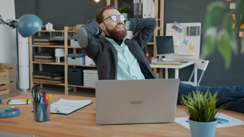 Portrait of happy guy relaxing in office sitting at desk smiling enjoying break and dreaming. Suceessful people, youth and creative workplace concept. Royalty-Free Stock Footage #1056391877