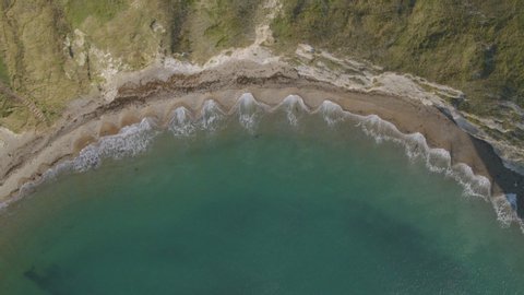 Top down, aerial drone shot of Man of War beach on the Jurassic Coast in Dorset, UK. Waves form patterns as they crash up on the perfectly curved bay.
