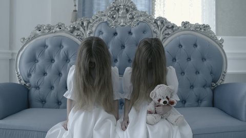 Creepy twins staring at cam sitting on old sofa with bloody toy in hands, zombie