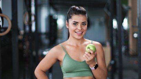 Portrait of smiling mixed race athletic woman eating fresh apple at gym. Gorgeous happy fitness girl posing enjoying healthy lifestyle and dietary nutrition. Medium shot on RED camera