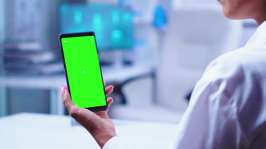 Medic in hospital cabinet holding phone with green screen wearing white coat while nurse opens glass door. Healthcare specialist in hospital cabinet using smartphone with mockup.