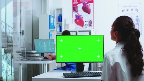 Doctor using computer with green screen mockup in hospital and assistant walks out of cabinet wearing blue uniform. Medic in white coat working on monitor with chroma key in clinic cabinet to check