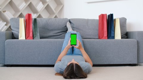 Online shopping, Buying from home, Credit card, Online store. A woman presses her finger on the smartphone screen while lying on the floor next to the sofa and shopping bags. Chromakey