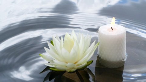 Spa background footage with water lily flower and large white candle. Slow motion of water splashing in pond with clouds reflection. Waterlily blooming in lake near stones underwater.