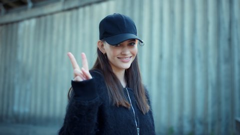 Serious woman showing v sign with fingers outdoors. Closeup smiling woman showing peace sign on urban street. Portrait of female protester showing victory gesture. Positive hipster looking at camera