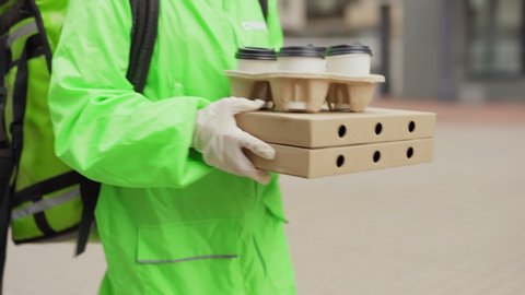 Tilt up tracking shot of young female food courier in safety face mask, gloves and green uniform carrying pizza boxes and takeaway coffee cups in her hands delivering order