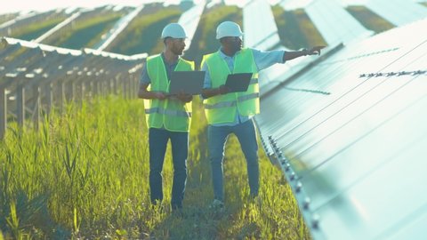 Close up engineers use laptop and project plan on the solar farm in special uniform standing look around discuss the installation of sunny batteries electricity environmental slow motion