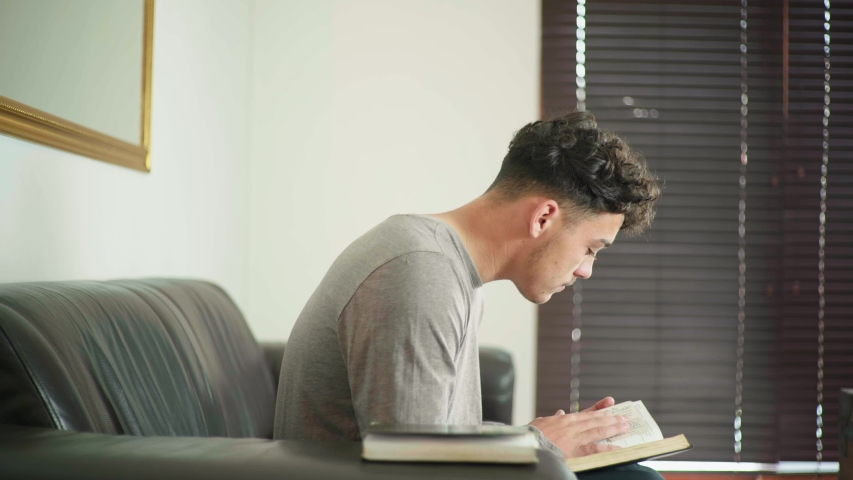 Mixed race guy sitting on couch reading the bible then closes the bible and starts praying. Royalty-Free Stock Footage #1056414551