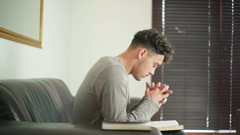 Mixed race guy sitting on couch reading the bible then closes the bible and starts praying.