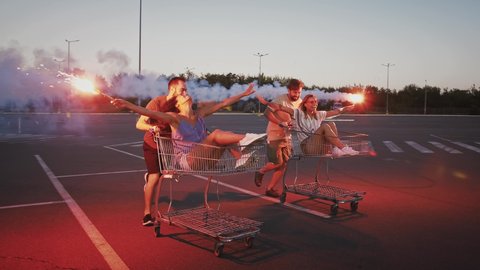 Guys riding and spinning on shopping trolleys with their girlfriends at deserted parking lot. Holding burning signal flares and laughing. Slow motion