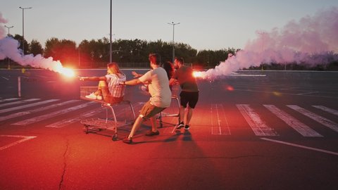 Multiethnic friends racing and spinning on shopping carts at deserted parking lot. Holding burning signal flares and laughing. Slow motion