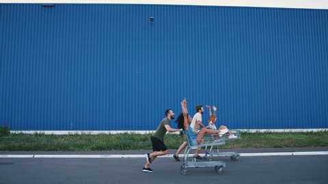 Young guys are racing on shopping carts with their girlfriends against blue wall of supermarket. Having fun and laughing. Slow motion