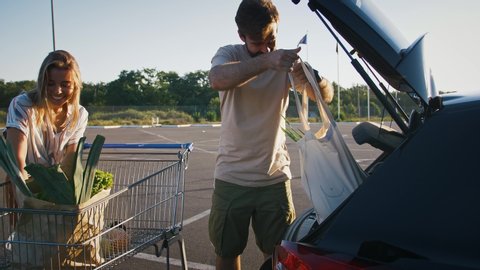 Girl and guy smiling, taking groceries out of cart and putting it into car. Parking lot, shopping in supermarket. Sunny morning. Eco bags, zero waste