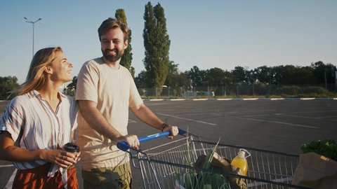 Young couple smiling and talking, walking by parking lot with cart of groceries after shopping in supermarket. Eco bags, zero waste. Slow motion