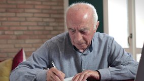 A senior man sitting at a table and using a laptop, taking some notes, at home. Slow camera movement.