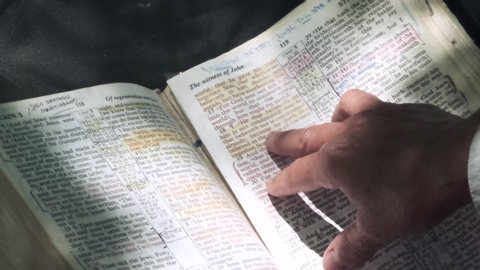 Adult Caucasian woman's finger points to and reads biblical scripture in gospel of John with sunlight shining on bible page with red letters, static close up