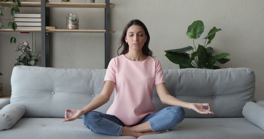 Mixed race Indian woman sit cross-legged on comfy sofa in living room closed eyes do meditation practice feels inner harmony and balance, improve self-conscious, mindfulness state, no stress concept Royalty-Free Stock Footage #1056430088