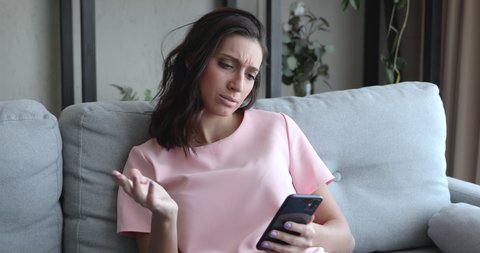 Indian mixed race woman sit on couch read message on phone feels upset, bad news information received, problems with device, connection lost, charging failure battery problem, new app not downloading