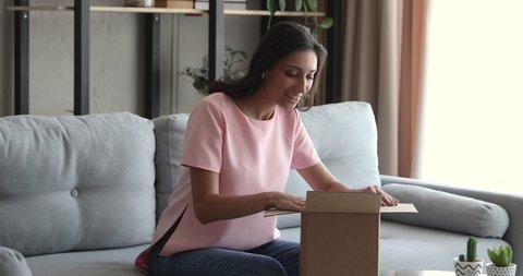 Indian ethnicity woman sit on sofa at home open post package shopping online bought goods on internet, happy young arab customer unpack postal shipping parcel feel satisfied by delivered order concept