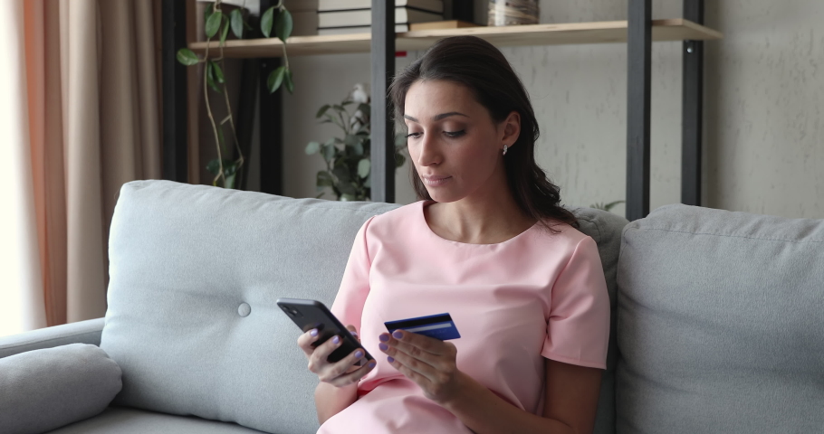 Middle eastern ethnicity young woman sitting on couch use credit card holding smartphone makes purchase smiles feels satisfied. Easy quick money transfer to relatives, ecommerce website user concept Royalty-Free Stock Footage #1056430205