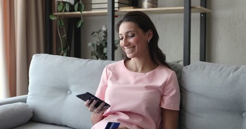 Middle eastern ethnicity young woman sitting on couch use credit card holding smartphone makes purchase smiles feels satisfied. Easy quick money transfer to relatives, ecommerce website user concept