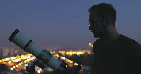 Astronomer with a telescope watching at the night sky with blurred city lights in the background.	