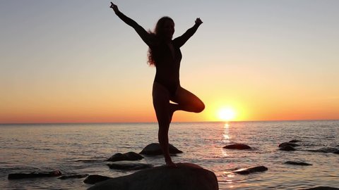 A young long-haired girl making yoga practice standing on a rock by the sea. Summer, sunset. Woman's silhouette