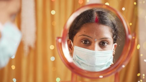 Indian woman getting ready infront of mirror by adjusting medical mask and sindoor or bindi during Diwali festival celebration - concept of new normal due to coronavirus or covid-19 pandemic