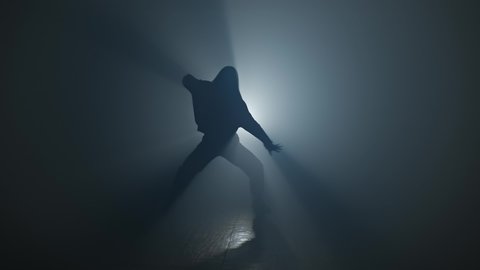 Silhouette of woman in hoody performing hip hop moves. Graceful female dancing in studio with smoke and spotlight. 4K, UHD