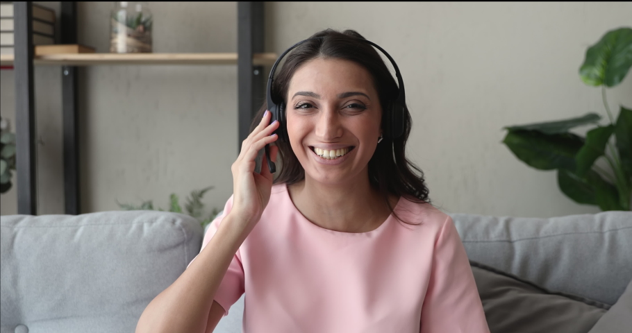 Head shot indian woman in headset sit on couch look at camera start conversation via video call. Webinar on-line training, candidate pass job interview remotely, modern tech easy comfort usage concept Royalty-Free Stock Footage #1056440234