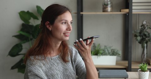 Woman sitting at workplace desk holding smartphone talking to friend on speaker phone, use phone app leave voicemail response, send voice message to colleague chatting on-line using wifi connection
