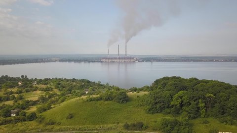Aerial view of small village between green trees and big lake with high chimney pipes polluting grey smoke from coal power plant.