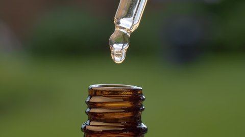 Macro shot essential oil dripping from a pipette on blurry natural background. Extreme close up hand taking transparent oil from a brown glass bottle