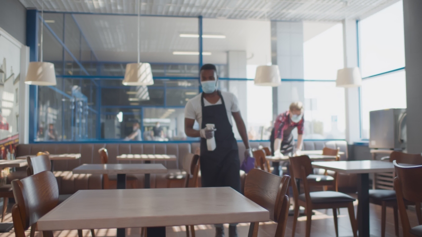 African male worker cleaning table with disinfectant in restaurant during coronavirus outbreak. Waiter in protective mask and gloves disinfecting table with spray and cloth Royalty-Free Stock Footage #1056442013