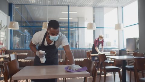 African male worker cleaning table with disinfectant in restaurant during coronavirus outbreak. Waiter in protective mask and gloves disinfecting table with spray and cloth
