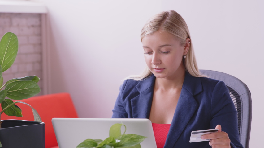 Smiling young caucasian woman customer holding credit card at laptop sitting at desk at home. Happy female shopper using instant easy mobile payments making purchase in online store. E-banking app | Shutterstock HD Video #1056448022