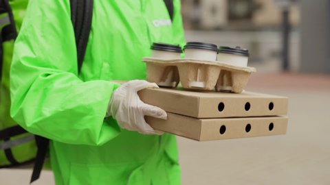 Tilt up tracking slow motion shot of female food courier in safety face mask, gloves and uniform carrying pizza boxes and takeaway coffee cups delivering order, thermal bag behind her back