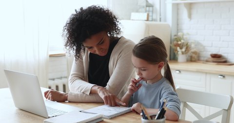 Adult afro american foster parent or babysitter helping little pretty caucasian girl preparing homework indoors. Caring biracial tutor giving private educational lesson to small attentive schoolgirl.