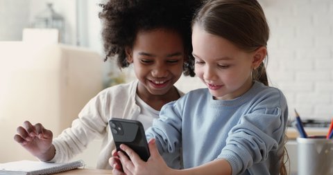 Head shot happy small mixed race friends playing online mobile games or using funny apps on smartphone together indoors. Addicted to tech diverse little children foster sisters having fun at home.