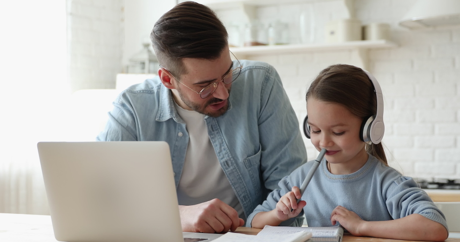Caring young father helping little primary school learner daughter doing homework, e-learning indoors. Smiling parent explaining education program to small girl in headphones, online study concept. Royalty-Free Stock Footage #1056450464